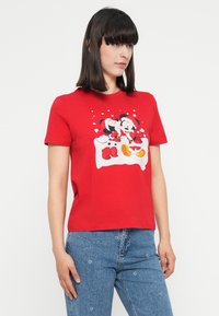 Woman wearing a red t-shirt with a festive Mickey and Minnie Mouse print and blue jeans with small white flower patterns.