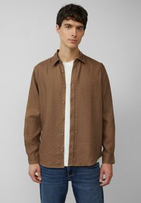 Chemise à manches longues en lin marron avec un col boutonné, dotée d'une patte de boutonnage et de détails en boutons, superposée à une chemise blanche.