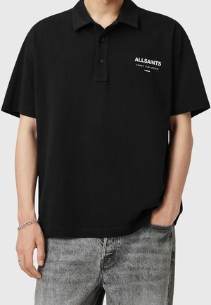 Person, der bærer en sort kortærmet polo-shirt med "ALLSAINTS" hvidt logo på brystet, parret med falmede grå jeans og et sølvarmbånd.
