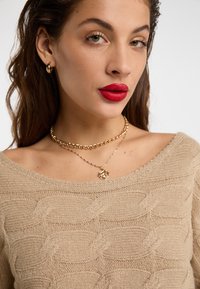 Pulover bej din tricot cu un model texturat în valuri, design off-shoulder și lanțuri de aur stratificate cu un pandantiv. Buze roșii cu accente aurii.