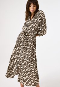 Knielange button-up jurk in een beige stof met een zwart geometrisch ruitpatroon, met lange mouwen en een strikdetail in de taille.