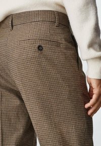 Ruskean ruudulliset housut, joissa on teksturoitu kangas, takatasku mustalla napilla, muotopoliisi ja vyölenkit.