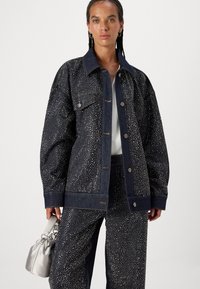 Chaqueta de mezclilla oscura con tachuelas metálicas, corte oversized, gran cuello, cierre de botones y bolsillos frontales, combinada con pantalones a juego con tachuelas y bolsa plateada.
