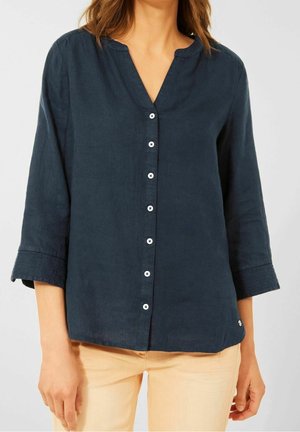 Woman wearing a dark blue button-up blouse with three-quarter sleeves and beige pants, standing against a plain background.