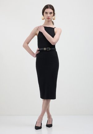 Abito midi aderente nero con design a spalla sola, caratterizzato da una ampia cintura nera con fibbia dorata. Il modello indossa tacchi a punta neri.
