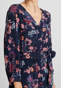 Blommönstrad klänning i marinblått med prickigt mönster. Har V-hals, långa ärmar, midja med snörning och varierande blommönster.