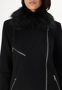 Černý kabát s límcem z umělé kožešiny a stříbrnými zipy vpředu. Tkanina má hladkou strukturu a kabát je střižený na tělo.
