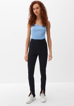 Blaues geripptes Tanktop mit schmalen Trägern, kombiniert mit dunkelblauen hochgeschnittenen Hosen mit seitlichen Schlitzen und weißen Turnschuhen.