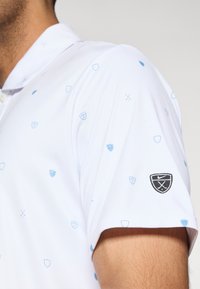 Λευκή μπλούζα πόλο με ανοιχτό μπλε γεωμετρικά σχέδια, κοντά μανίκια και μαύρο λογότυπο Nike με σταυρωμένα κλαμπ στο δεξί μανί κι.