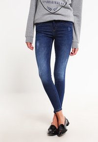 Jeans skinny de color azul oscuro con áreas desvaídas y un sutil efecto de desgaste, combinados con mocasines negros de charol. Material de mezclilla con una textura suave.