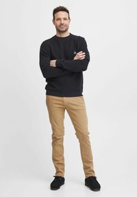Felpa in cotone nera con collo a giro e dettaglio del logo, abbinata a chinos beige e scarpe nere. Mostrata su uno sfondo neutro.