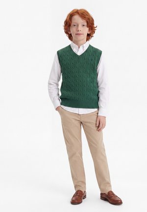 Jongentje met rood haar die een groene gebreide vest draagt over een wit overhemd, beige broek en bruine leren loafers, staande met één hand in de zak.