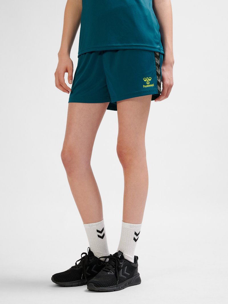 Person trägt türkisfarbene Sportshorts und passendes Oberteil, weiße Socken mit schwarzem Chevron-Muster und schwarze Laufschuhe, steht auf grauem Boden.