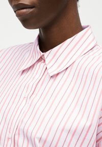 Mørk hudfarve person iført en lyserød og hvid vertikalt striped skjorte med knapper og spids krave, uden synlige accessories.