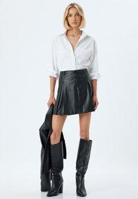 Schwarzer Lederrock mit Falten, kombiniert mit einem weißen Hemd. Modell trägt lange schwarze Lederstiefel und hält eine passende Jacke.