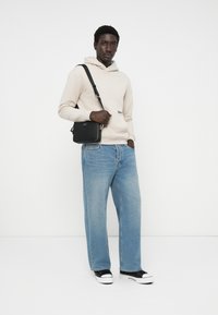 Felpa beige con cappuccio, jeans a gamba larga blu, borsa a tracolla nera e sneaker nere con suole bianche su uno sfondo semplice.