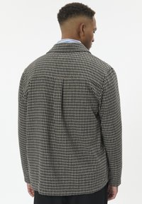 Mand vender væk iført en sort og beige ternet jakke over en blå skjorte med krave, stående mod en ensfarvet hvid baggrund.