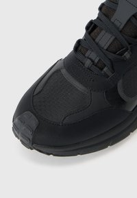 Zapatilla deportiva negra con un patrón hexagonal texturizado, materiales sintéticos lisos, puntera reforzada y cordones negros.