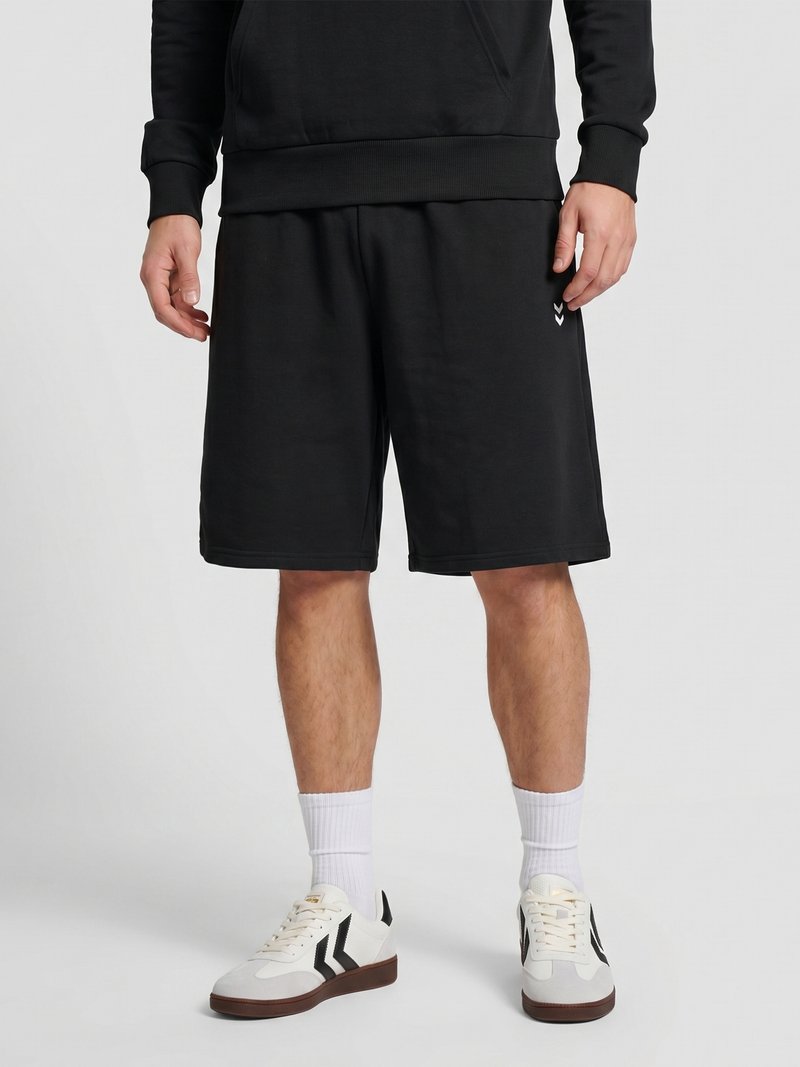 Man wearing black knee-length shorts, white socks, white sneakers with black accents, and a black sweatshirt standing against a plain background.