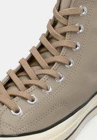 Sneakers înaltă din piele bej cu suprafață texturată, șireturi plate bej, ochiuri argintii și o talpă albă contrastantă.