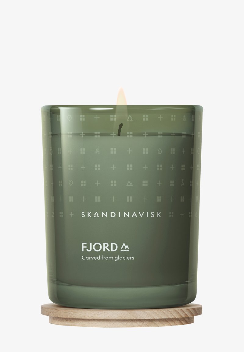 Bougie en verre de couleur vert muet avec des symboles gravés ; marquée "FJORD" et "Taillée dans des glaciers," posée sur un sous-verre en bois clair. Flamme visible.
