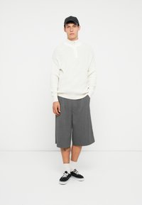 Λευκό ριγωτό πουλόβερ με ψηλό λαιμό, συνδυασμένο με γκρι πλισέ σορτς. Φορεμένο με μαύρα αθλητικά παπούτσια και λευκές κάλτσες. Το φόντο είναι απλό.