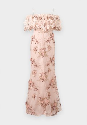 Femme portant une robe longue rose poudré avec un corsage à épaules dénudées orné d'applications florales et de fleurs brodées, debout devant un fond clair uni.