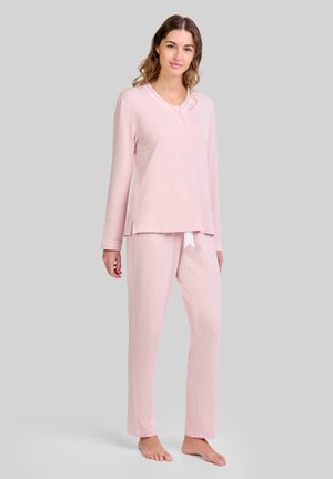 Joven mujer de pie descalza, vestida con una blusa de manga larga de color rosa claro con botones y pantalones a juego, frente a un fondo liso.