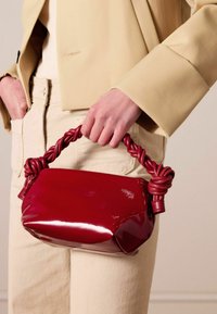 Red patent leather handbag with a knotted red strap, rectangular shape, glossy texture, and minimalistic design.