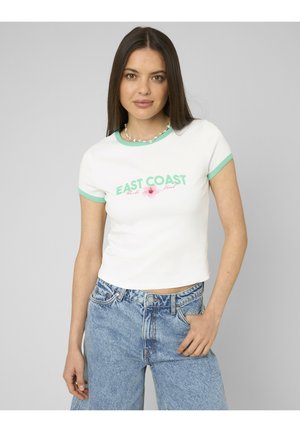 Femme aux longs cheveux bruns portant un t-shirt blanc ajusté avec une bordure verte et le texte « EAST COAST », associé à un jean taille haute bleu clair.