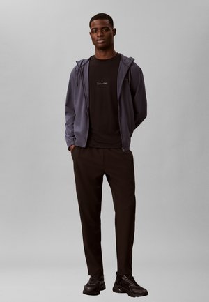 Mann trägt ein schwarzes Calvin Klein T-Shirt, eine graue Kapuzenjacke mit Reißverschluss, schwarze Hosen und schwarze Sneaker und steht mit den Händen in den Taschen.
