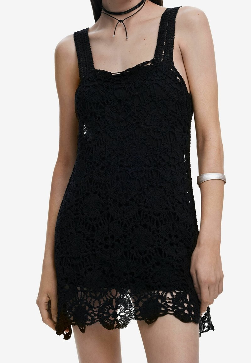 Robe noire sans manches en crochet avec des motifs floraux, portée avec un collier ras du cou noir fin et un bracelet argenté au bras gauche.