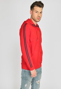 Rød zip-up hoodie lavet af blødt stof, med marineblå striber langs ærmerne. Designs inkluderer en frontlomme og justerbar snørredragt.