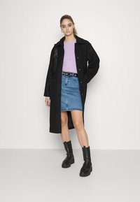 Casaco preto em mistura de lã com botões, combinado com uma blusa lilás clara e uma saia de ganga azul. Botas de tornozelo pretas completam o conjunto.