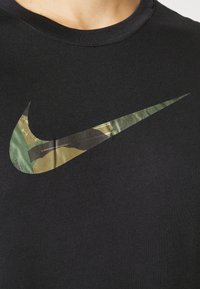 Camiseta de algodón negra con un gran logotipo de Nike en forma de swoosh con patrón de camuflaje en el frente. Cuello redondeado, textura lisa y mangas cortas.
