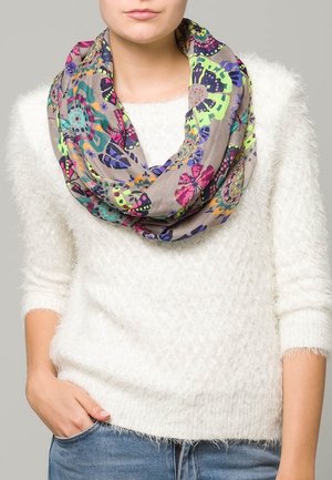 Personne portant un pull blanc texturé et une écharpe infinie à motif floral coloré, les mains dans les poches d'un jean bleu.