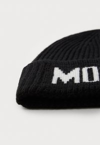 Zwarte gebreide beanie met ribbelstructuur. Witte letters staan weergegeven op de omgeslagen rand, wat in contrast staat met het donkere materiaal.
