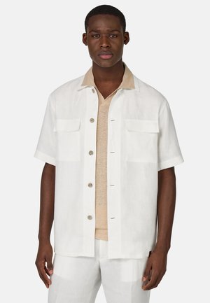 Camisa blanca de manga corta abotonada con dos bolsillos en el pecho, combinada con una camiseta de cuello en V de color beige. La tela parece ligera y texturizada.