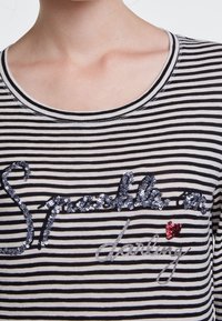 Raidallinen paita mustassa ja kermassa, jossa on pyöreä pääntie ja koristeellinen paljettikirjoitus, joka sanoo "Sparks a darling" punaisella sydämen yksityiskohdalla.