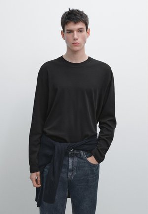 Jonge man met kort donker haar, gekleed in een zwart shirt met lange mouwen, donkere spijkerbroek en een donker vest om zijn middel geknoopt, staand tegen een effen achtergrond.