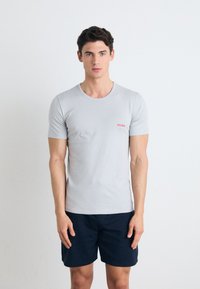 Lys grå bomulls T-skjorte med korte ermer og rund hals, med en liten rød "HUGO" logo på venstre bryst. Bært med marineblå shorts.