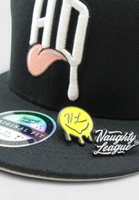 Musta musta baseballi hattu, jossa on valkoista brodeerattua tekstiä, vaaleanpunainen kielipisto ja kaksi nappia: keltainen valuva hymyilevänaama ja musta "Naughty League" -logo.