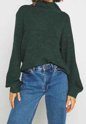 Frau trägt einen dunkelgrünen grob gestrickten Pullover mit Ballonärmeln, der in eine hoch taillierte blaue Jeans gesteckt ist, vor einem schlichten Hintergrund.