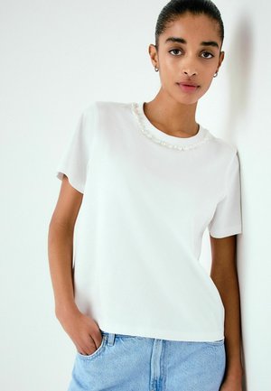 Vrouw met kort zwart haar, gekleed in een wit blouse met korte mouwen en een met kralen versierde halslijn, en lichtblauwe spijkerbroek, staand tegen een witte muur.