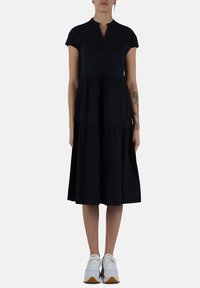 Abito nero in cotone con maniche corte, scollatura a V e gonna a balze arricciate. Abbinato a sneakers bianche.