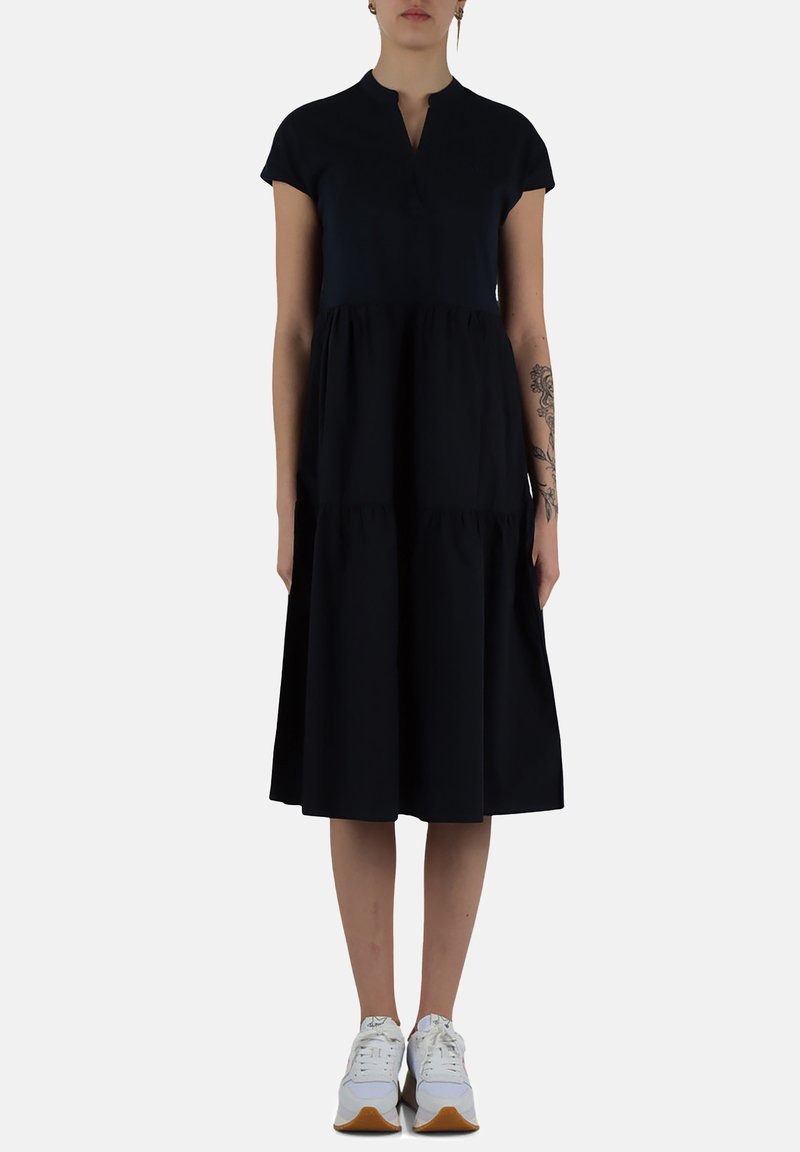 Abito nero in cotone con maniche corte, scollatura a V e gonna a balze arricciate. Abbinato a sneakers bianche.