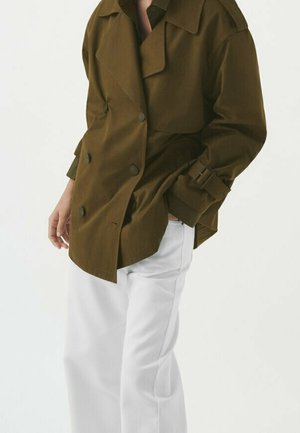 Veste double boutonnage vert olive avec un grand col, une ceinture latérale et un tissu texturé, associée à un pantalon blanc à jambes droites.