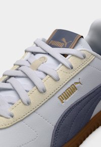 Λευκό δερμάτινο αθλητικό παπούτσι με ανοιχτό κίτρινα και γκρι στοιχεία, διαθέτει επίπεδα λευκά κορδόνια, καφέ-χρυσό λογότυπο και υφασμένο ελαστικό πέλμα.