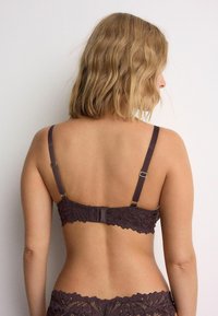 Pruun pitsrinnahoidja reguleeritavate rihmade, konkslukkude ja lainelise servaga. Riide tekstuuril on peen lillemuster.