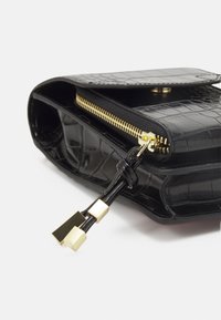 Black crocodile-embossed leather bag with a zip closure, gold zipper hardware, and a cord with square gold accents. Textured surface.